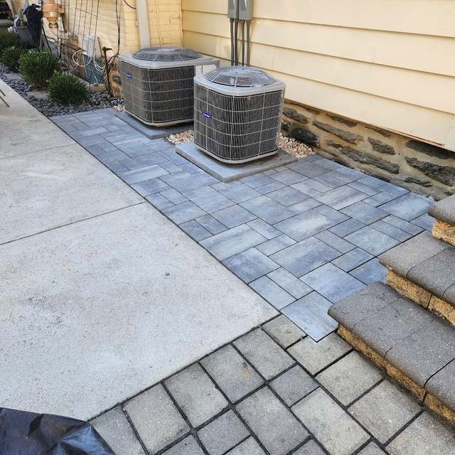 A new gray paver pad around two air-conditioning condensers, meeting a concrete walk and a set of paver steps along the side of a house.