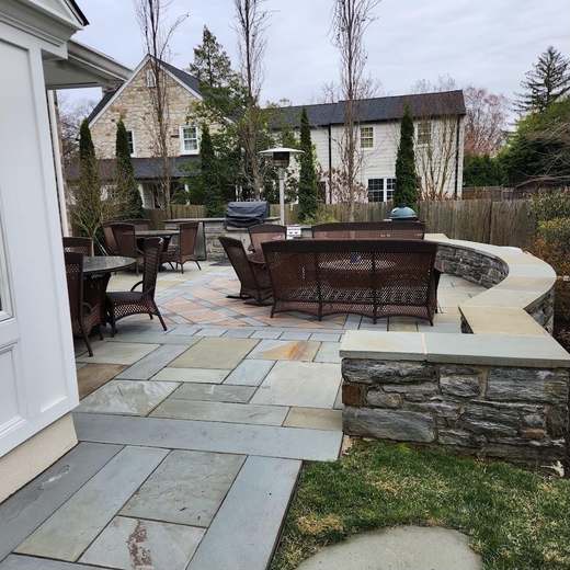 A bluestone patio with a curved stone seat wall capped in bluestone, set with outdoor dining and lounge furniture beside a house.