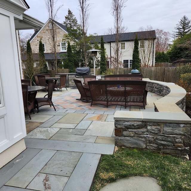 A bluestone patio with a curved stone seat wall capped in bluestone, set with outdoor dining and lounge furniture beside a house.