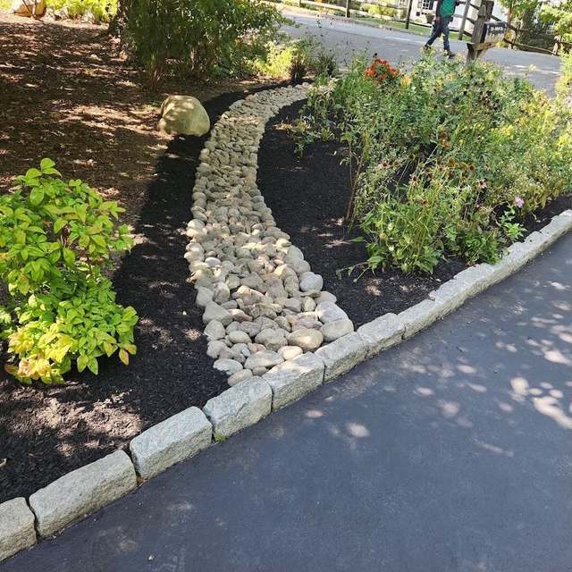 A dry creek bed of river stone curving through black mulch to a granite cobble curb at the edge of a driveway, with perennials in bloom beside it.