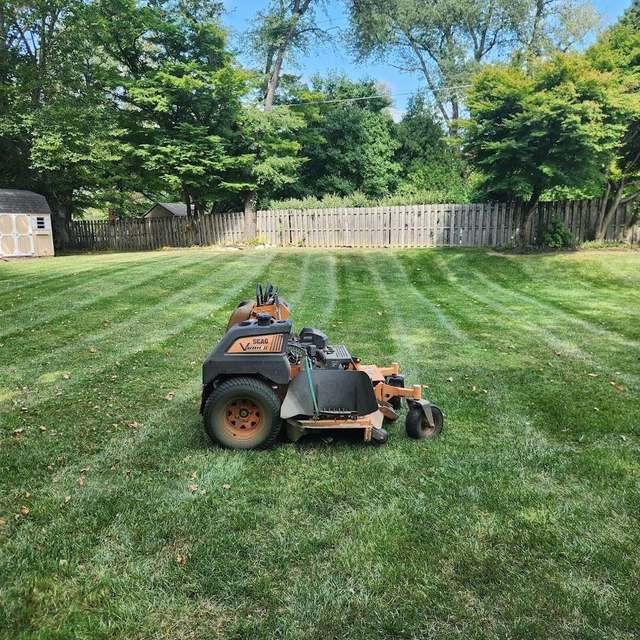 A stand-on mower parked on a rear lawn cut in clean diagonal stripes, with a wood fence and trees behind.
