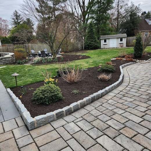 A curved paver walk bordered in granite cobble, wrapping a mulched bed, with a lawn, a fire pit seating area and a shed beyond.