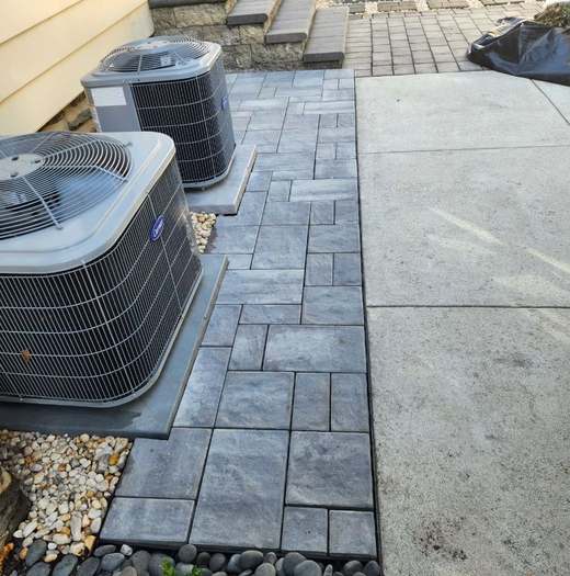 A grey paver pad and walkway laid in a running bond with a soldier-course border, meeting an existing concrete walk and stone steps.