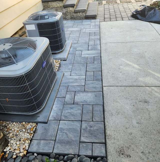 A grey paver pad and walkway laid in a running bond with a soldier-course border, meeting an existing concrete walk and stone steps.