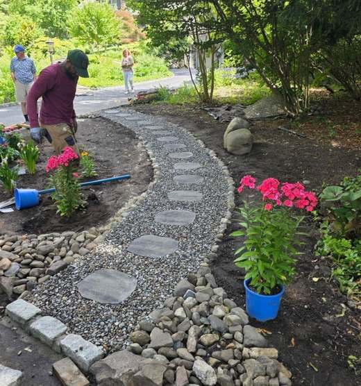 Two crew members planting along a new gravel path, with flats of perennials and a wheelbarrow on the open ground.