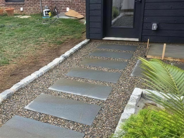 Large stone stepping slabs set in pea gravel with a granite cobble edge, leading to the side door of a house clad in black siding.